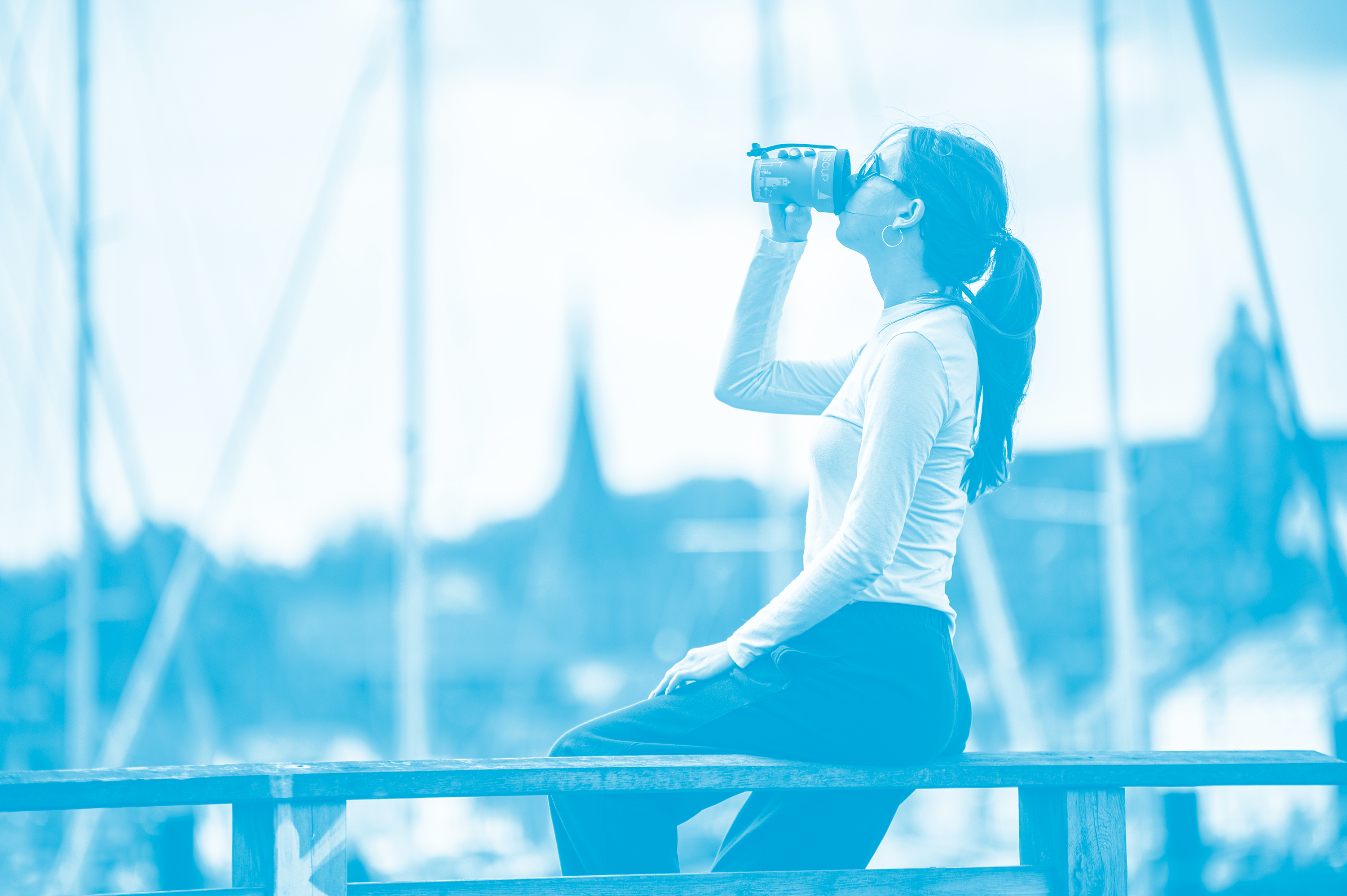 Eine Frau sitzt am Flensburger Hafen und trinkt aus dem FLENSCUP; im Hintergrund ist die Flensburger Innenstadt zu sehen.